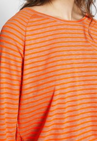 Orange randig långärmad topp med smala och breda ränder, vid halsringning och mjukt material. Tyget verkar vara lätt och andningsbart.