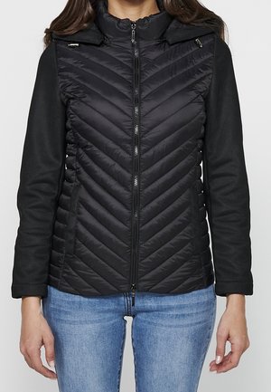 Mujer que lleva una chaqueta negra acolchada con cremallera, costura en forma de espiga y mangas negras sólidas, combinada con vaqueros azul claro.