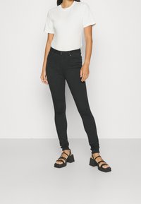 Vaqueros skinny de mezclilla negra de cintura alta, combinados con una camiseta blanca y sandalias negras con tiras que tienen un tacón grueso.