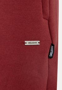 Rødt bomuldsblandet stof med en tekstureret overflade. Indeholder et sølvfarvet "SMILODOX" mærke og en sort label med detaljer på siden.