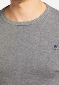 Pull gris en maille avec un col côtelé, arborant un petit logo de crâne bleu sur le côté gauche de la poitrine. Texture douce, coupe ajustée.