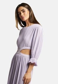 Lavendelfarbenes Plissee-Kleid mit langen Puffärmeln, Ausschnitt in der Taille und Rüschensäumen. Glatte Stofftextur und fließende Silhouette.