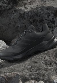 Scarpa sportiva nera con suola spessa e lacci decorati che riposa su un terreno accidentato e roccioso.