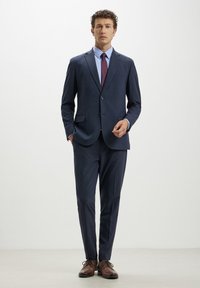Completo blu navy con giacca a chiusura a un bottone e due tasche frontali, abbinato a pantaloni slim-fit e scarpe eleganti marroni.