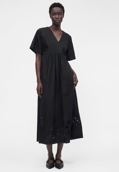 Femme portant une robe mi-longue noire à col en V, manches courtes et détails floraux découpés à l'ourlet, associée à des ballerines noires pointues.