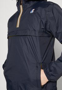 Cortavientos azul marino con cremallera frontal de medio cierre, puños elásticos y detalles contrastantes en amarillo y blanco. Incluye un bolsillo tipo canguro y un logo de la marca.