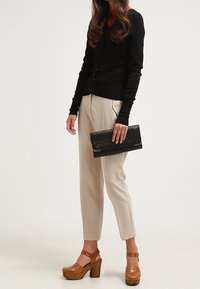 Svart långärmad cardigan, beige tapered byxor med framseams, bruna plattformsskor och en svart clutch med en texturerad design.