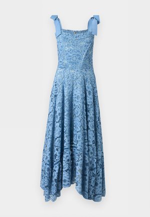 Γυναίκα που φοράει μπλε δαντελωτό φόρεμα με ανθισμένα μοτίβα και δεσίματα στους ώμους, στέκεται με ένα χέρι στη μέση και λευκές μυτερές γόβες.