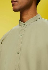 Gros plan d'une personne portant une chemise beige clair boutonnée avec un col mandarin sur un fond jaune.