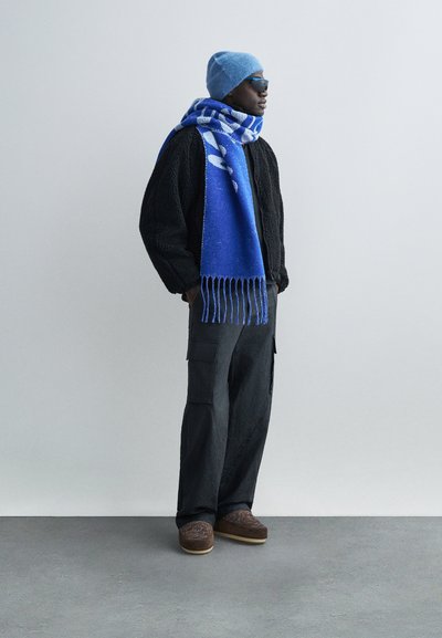 Bărbat purtând o căciulă tricotată albastră, eșarfă albastră cu model, geacă neagră din fleece, pantaloni cargo închiși la culoare și pantofi maro brodați, stând pe un fundal simplu.