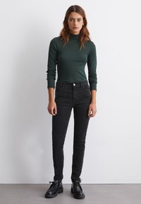 Mustad kõrgema tooniga mustad skinny teksad, millel on kerge venitus, on kombineeritud tumeroheline fikseeritud kõrge kaelusega särk. Mudel seisab käed külgedel.