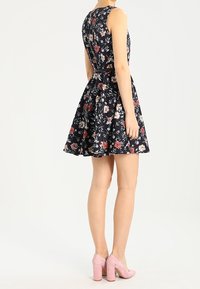 Blommig klänning i marinblått med figursydd överdel och utsvängd kjol; har dragkedja i ryggen och bälte. Matchas med rosa högklackade skor.