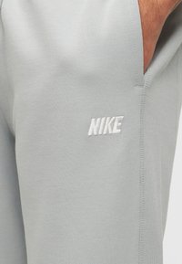 Κοντινό πλάνο από τσέπη ανοιχτόγκρι φορμας Nike με χέρι μέσα και λευκό κεντημένο λογότυπο Nike κοντά στη ραφή.