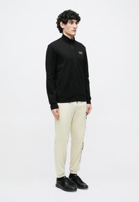 Čierna bunda na zips s vysokým golierom a zlatým logom, skombinovaná s krémovými teplákmi s potlačou na boku a čiernymi teniskami.