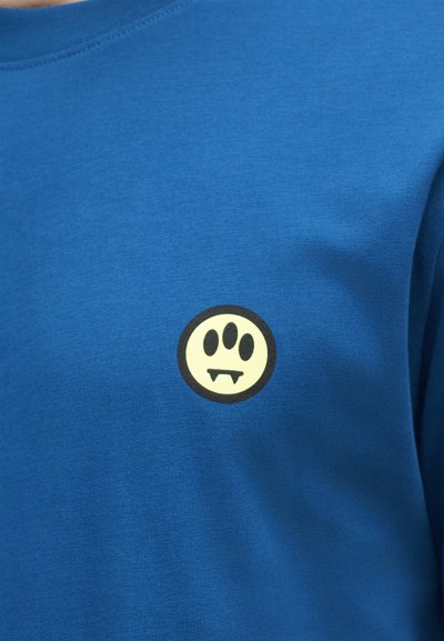 Μπλε πουκάμισο με ένα μικρό στρογγυλό λογότυπο που δείχνει ένα κίτρινο πρόσωπο με τρία μαύρα μάτια και βαμπιρικά δόντια στο κέντρο.