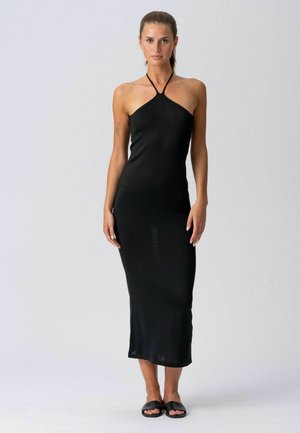 Nő áll egy fekete ujjatlan halter midi ruhában és fekete papucsban, egyszerű világos háttér előtt.