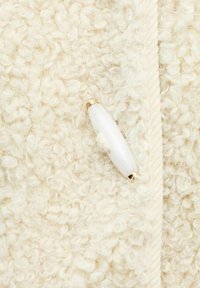 Tessuto texture color crema con fibre ricce, dotato di una chiusura liscia bianca e dorata. Il tessuto presenta un aspetto morbido e soffice.