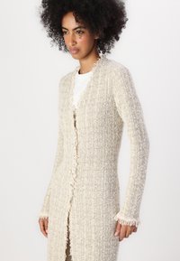 Long gilet ouvert en beige, présentant un motif de tricot texturé, des détails à franges le long des bords et des manches ajustées.