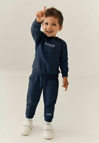 Dunkelblaue Sweatshirt und passende Hose mit einem weißen "BAKER"-Logo. Weiße Sneakers mit Klettverschlüssen runden das Outfit ab.