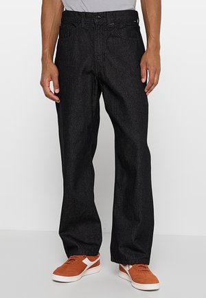 Mand iført løstsiddende sorte denimjeans og brune sneakers med hvide detaljer, stående mod en ensfarvet hvid baggrund.
