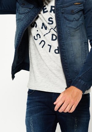 Giacca di jeans blu con due tasche sul petto, chiusura con zip e texture sbiadita. Indossata sopra una maglietta grigia chiara con bold testo circolare nero.