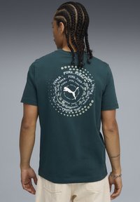 Tmavě zelené bavlněné tričko s kruhovou grafikou, na které je slovo "PUMA" v různých velikostech a siluetou loga, umístěné na tečkovaném pozadí.