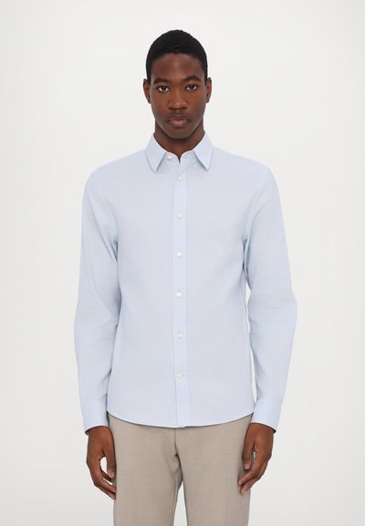 Camisa de botón de manga larga en azul claro, con un cuello clásico, presenta botones blancos y una textura de algodón suave. Diseño sencillo, sin patrones.