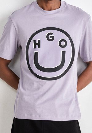 Person wearing a light purple short-sleeve shirt with a black and white circular smiley face design and the word "HUGO" inside.
