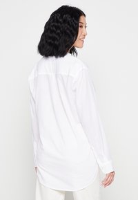 Camicia bianca a maniche lunghe realizzata in tessuto leggero, con orlo arrotondato, polsini con bottoni e dettaglio della cucitura del girocollo sul retro.