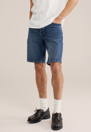 Mees, kellel on seljas sinised teksapüksid, valged sokid, mustad paeltega kingad ja valge T-särk, üks käsi taskus.