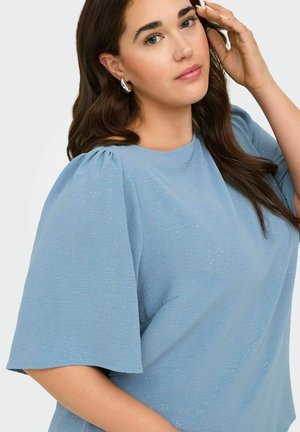 Mujer con cabello largo y castaño que lleva una camiseta de manga corta azul claro con sutiles destellos y pendientes de aro plateados, tocándose la cara.