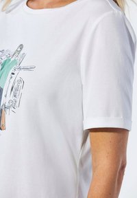 Weißes Baumwoll-T-Shirt mit kurzen, aufgerollten Ärmeln. Besitzt ein grafisches Design auf der Vorderseite einer Frau in einem grünen Kleid, was einen lässigen Look betont.