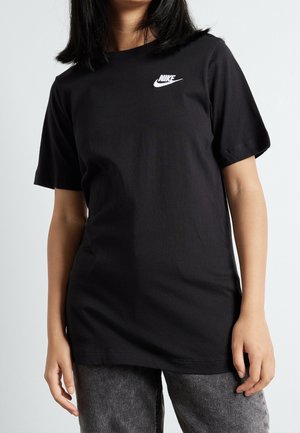 Person iført en sort Nike T-shirt med et hvidt logo på venstre bryst, kombineret med grå jeans mod en ensfarvet lys baggrund.