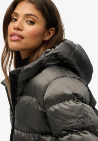 Veste noire rembourrée avec un col haut et une capuche à cordon de serrage, dotée d'une fermeture éclair et d'un logo "SUPERDRY" brodé sur la manche.