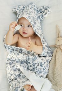 Huppupyyhkeä vauvalle valkoisena sinisellä kukkakuviolla, jossa on pupunkorvat. Vauva pitää tuttia kädessään istuessaan tekstuurilla varustetulla peitolla.