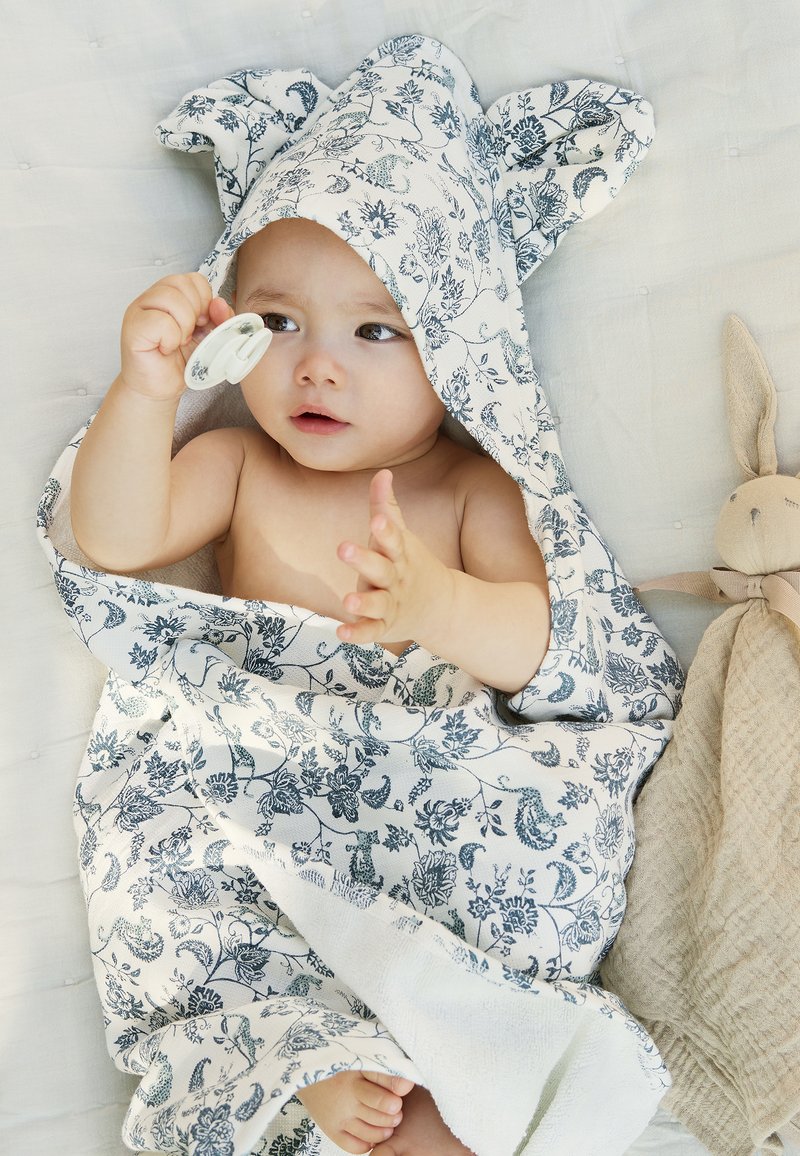 Huppupyyhkeä vauvalle valkoisena sinisellä kukkakuviolla, jossa on pupunkorvat. Vauva pitää tuttia kädessään istuessaan tekstuurilla varustetulla peitolla.