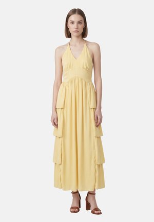 Femme portant une robe maxi jaune pâle sans manches à col licou avec des panneaux latéraux superposés et des sandales marron à talons carrés et orteils ouverts.