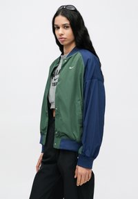 Ανδρικό bomber μπουφάν με χρωματικό σχέδιο σε πράσινο και ναυτικό μπλε. Ελαφρύ υλικό, εμπρόσθιο κλείσιμο με κουμπιά, ελαστικά μανίκια και ζώνη στη μέση.