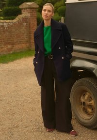 Námornícky dvojradový kabát so striebornými gombíkmi, zelený top, čierne široké nohavice a vínové topánky. Pozadie tvorí štrk a pneumatiky vozidla.