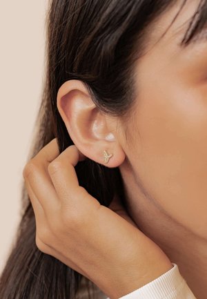 Gold stud earrings in a butterfly shape with small sparkling accents. A hand is adjusting hair near the ear, showing the earring's placement.