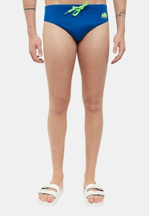 Uomo che indossa slip da bagno blu con coulisse verde e sandali bianchi, vista frontale dalla vita ai piedi su sfondo bianco.