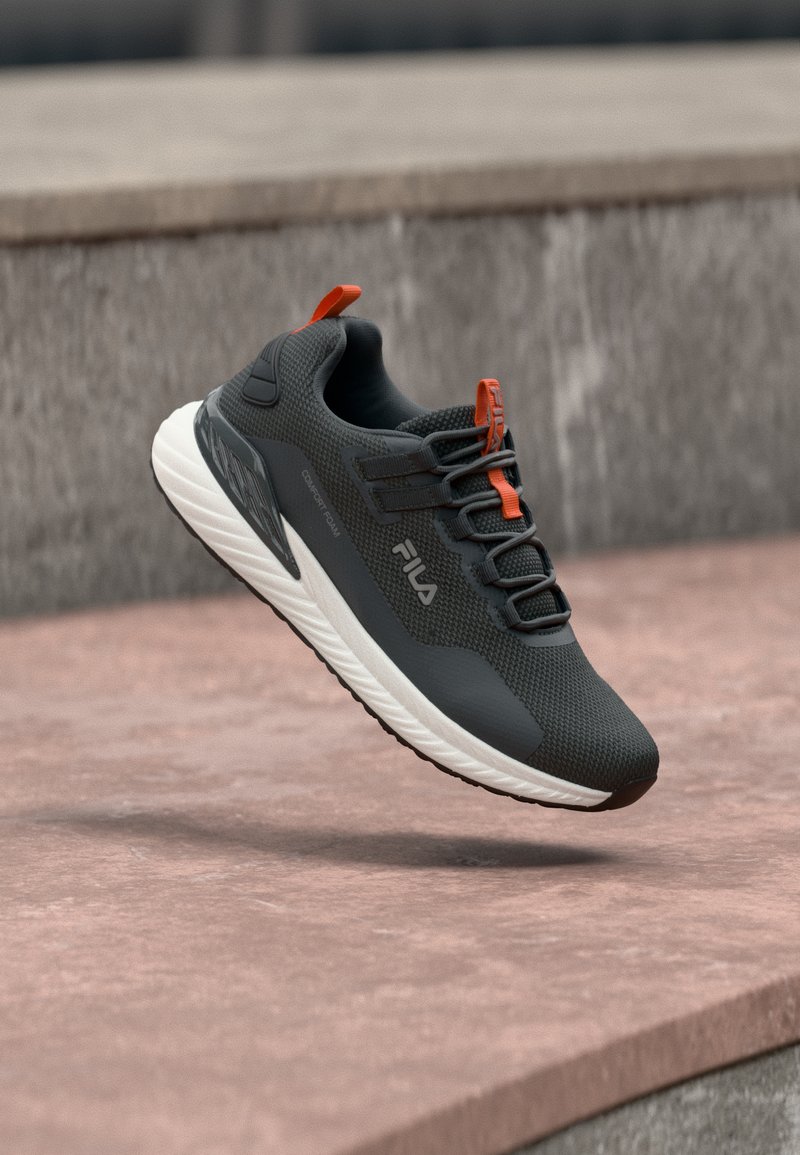 Chaussure de sport Fila gris foncé et orange avec une semelle blanche, affichée en plein air au-dessus d'une surface en béton marron.