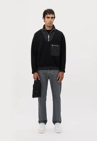 Felpa nera con zip a mezzo busto, tasca frontale e dettagli grigi, abbinata a jeans grigi e sneakers bianche. Modella che tiene una borsa nera.