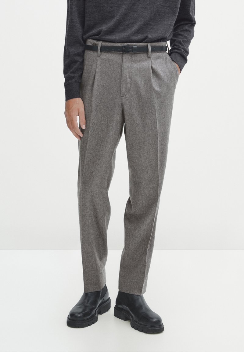 Pantalon en laine grise à coupe décontractée, avec plis sur le devant, ceinture, et bottines noires. Le tissu présente une texture subtile.