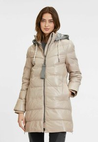 Cappotto puffer beige con texture lucida, maniche lunghe, cappuccio grigio e chiusura frontale con zip; presenta tasche laterali e un motivo trapuntato orizzontale.