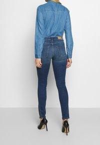 Vaqueros skinny de denim en azul oscuro, con cintura alta, dos bolsillos traseros y detalles de cremallera en los tobillos. Combinados con una camisa de color azul claro.