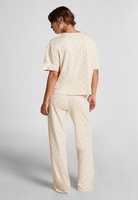 Woman with brown hair in a low bun wearing loose beige pajama set with subtle small pattern, standing and facing away against plain background.