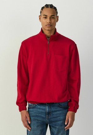 Young man with braided hair wearing a red quarter-zip pullover with a chest pocket and blue jeans standing against a plain background.