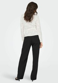 Blouse blanche à manches longues avec des motifs floraux et un bord en dentelle, associée à un pantalon noir à jambes droites et des talons noirs. Vue de dos.