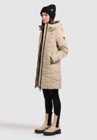Manteau doudoune beige matelassé avec capuche, doté de poches latérales et d'une fermeture éclair sur toute la longueur, associé à des leggings noirs et des bottines beiges.